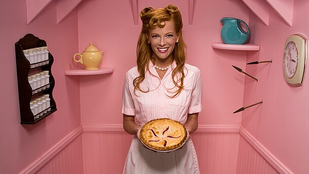 Eine Frau in einem rosafarbenen Kostüm und Perlenkette hält einen Kuchen in der Hand